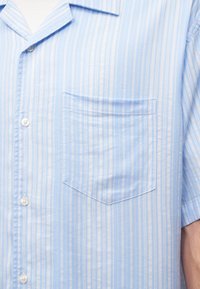 Chemise bleu clair à manches courtes avec des rayures verticales blanches, des boutons à l'avant et une poche poitrine à gauche.