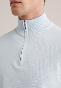Svetlo modra pulover z visokim ovratnikom in polovičnim zadrgo. Teksturirana tkanina, rebrasti ovratnik in gladek, oprijet videz.