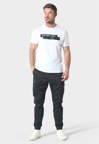 Witte t-shirt met zwarte grafische tekst, gecombineerd met zwarte cargobroek met zijzakken, afgewerkt met witte sneakers.