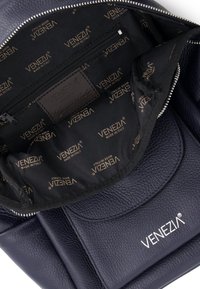 Marineblå læderrygsæk indvendigt med en lynlåslomme og glat stofforing, der har et gentaget "VENEZIA"-logo i guld.