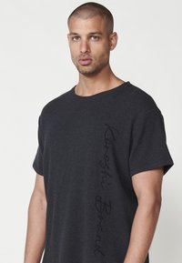 Tmav gray oversized tričko z textúrovanej tkaniny, s okrúhlym výstrihom a vyšívaným detailom "Kareshi Brand" po strane.