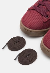 Červené tenisky z manšestru s hnědými gumovými podrážkami; vedle nich jsou dva hnědé kulaté tkaničky. Texturovaná tkanina a zaoblený tvar špičky.