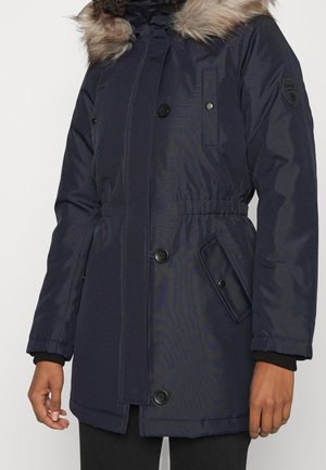 Navyblauwe winterparka met bont afgekapte capuchon, knoop- en drukknopsluitingen, elastische taille en zijflapzakken gedragen door persoon met donkere broek.