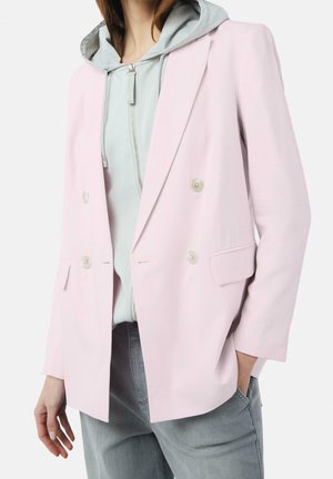 Persona que lleva un blazer cruzado rosa claro sobre una sudadera con capucha gris claro con cremallera y pantalones grises, con una mano en el bolsillo.