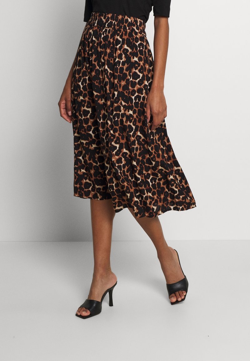 En kvinna står klädd i en svart topp och en knälång kjol med leopardsmönster, som är kombinerad med svarta klackskor. Bakgrunden är neutral.