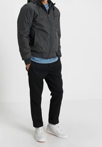 Giubbotto bomber grigio scuro con cappuccio, zip frontale e due tasche, abbinato a pantaloni neri con risvolto e sneakers bianche. Outfit casual.