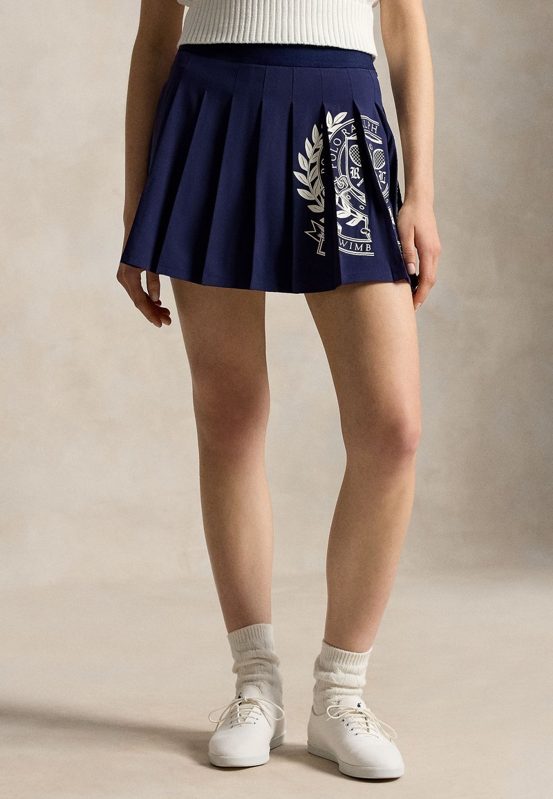 Woman wearing a navy blue pleated tennis skirt with white crest, white socks, white sneakers, and a white sleeveless top, standing.