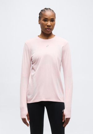 Naine punutud juustega, kandes heleroosa pika varrukaga spordipluusi ja musti retuuse ühevärvilise tausta ees.