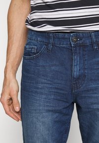 Jeans da uomo in denim blu scuro con una leggera scoloritura, dotati di chiusura a bottoni, tasche anteriori e tessuto texturizzato. Camicia a righe parzialmente visibile.