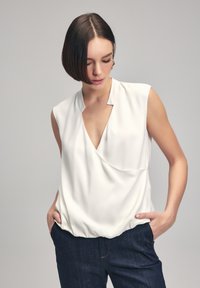 Blusa bianca senza maniche con scollatura a V, design drappeggiato e orlo elasticizzato, indossata con jeans in denim scuro. Tessuto liscio e leggero.