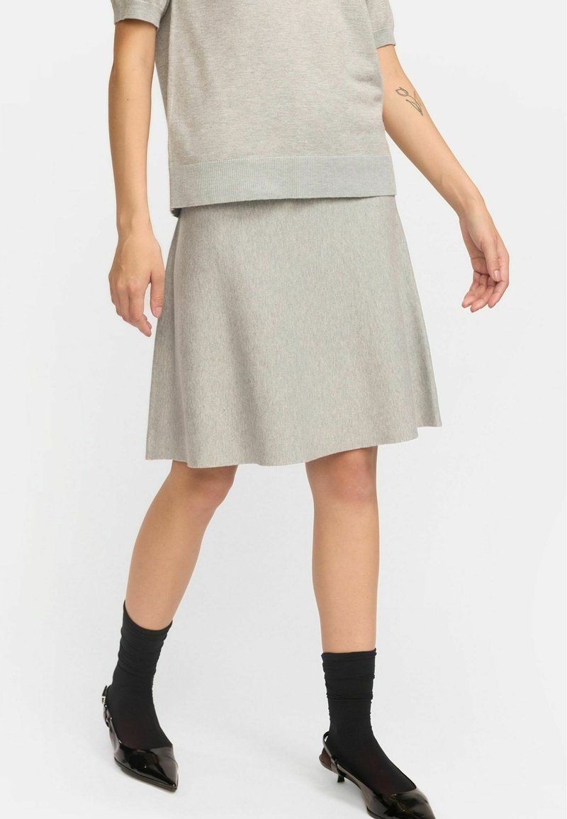 Jupe A-line grise claire, arrivant aux genoux, avec une texture lisse, associée à des chaussettes noires ajustées et des escarpins noirs à bout pointu brillants.