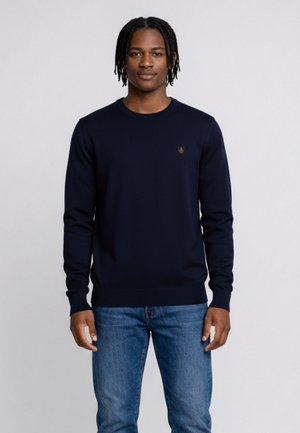 Jonge man met korte dreadlocks, gekleed in een marineblauwe trui met lange mouwen en een blauwe spijkerbroek, staand tegen een effen lichte achtergrond.