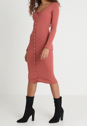 Femme portant une robe rose poudré boutonnée à manches longues, longueur genou, et des bottines noires à talons hauts, debout sur un sol blanc.