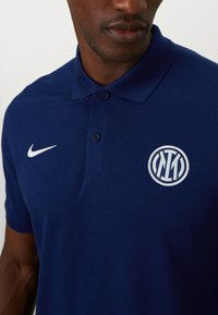 Mørkeblå polo tee lavet af tekstureret stof, med en to-knap placket, hvid Nike-logo og et broderet rundt logo på brystet.