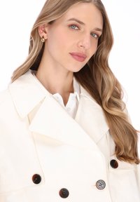 Weißer Trenchcoat mit großen Revers, braunen Knöpfen und glatter Textur. Das Modell trägt darunter ein weißes Hemd und goldene Tropfenohrringe.