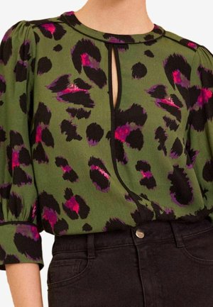 Grüne Bluse mit schwarzem und pinkem abstraktem Leopardenmuster, Schlitz am Ausschnitt, gepuffte 3/4-Ärmel, in hoch taillierte schwarze Jeans gesteckt.