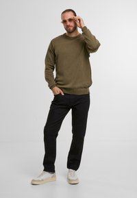 Maglione lavorato a maglia verde oliva con collo rotondo e polsini a costine, abbinato a pantaloni neri a gamba dritta e sneakers con dettagli beige.