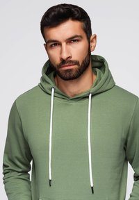 Sudadera verde con una tela texturizada, capucha con cordón en blanco contrastante y bolsillo canguro. Diseño simple y contemporáneo.