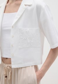 Weißes, gekürztes Shirt aus leichtem Stoff, mit kurzen Ärmeln und einem strukturierten Spitzen-Detail auf der Tasche. Knöpfe sind entlang der Vorderseite sichtbar.