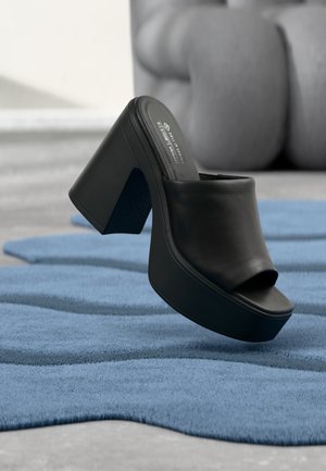 Zapato mule negro con plataforma, tacón alto y puntera abierta, apoyado sobre una alfombra azul texturizada con muebles grises desenfocados al fondo.