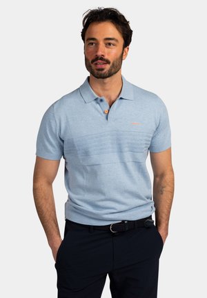 Man met baard, gekleed in een lichtblauw polo met korte mouwen en een zwarte broek, staat met handen in de zakken tegen een effen achtergrond.