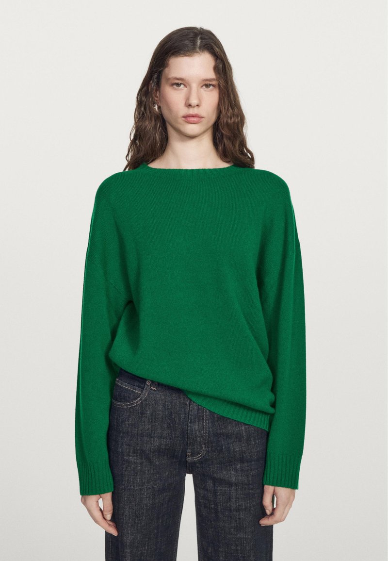Grüner Oversize-Pullover aus weichem Material mit rundem Ausschnitt und langen Ärmeln, kombiniert mit dunkelblauer Jeans.
