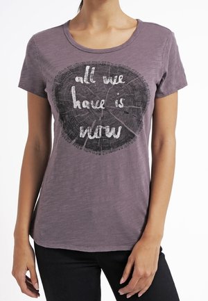 Frau trägt ein violettes Hemd mit einem Baumring-Design und dem Text "all we have is now" auf der Vorderseite.