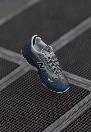 Donkerblauwe en grijze New Balance 1000 sneaker met witte veters, zwevend boven een metalen rooster vloer.