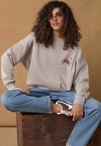 Hanorac oversized de culoare gri deschis, cu broderie florală, asortat cu blugi largi de culoare albastră și adidași maro. Așezat pe o suprafață din lemn.