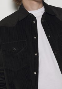 Camicia in corduroy nera con bottoni a pressione, due tasche sul petto e un tessuto testurizzato, indossata sopra una maglietta bianca semplice.