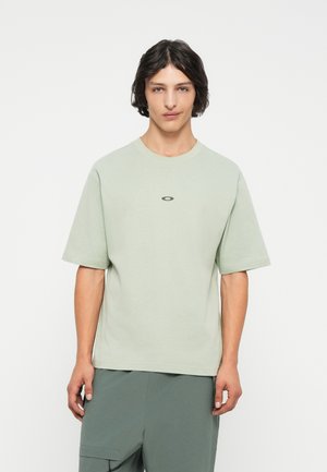 Tricou din bumbac verde deschis, cu guler rotund, mâneci scurte și un logo oval pe piept. Textură netedă; croi lejer.