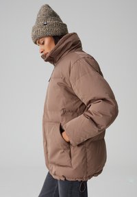 Veste matelassée isotherme marron avec un col montant, une fermeture éclair, des poches latérales et un ourlet élastique. Accompagnée d'un bonnet en tricot texturé.
