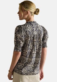 Sieviete ar blondiem matiem, kas sašūta bunā, ģērbusies tumši zila un baltā ziedu raksta puff-maki bluzē un bēšās apakšbiksēs, stāv ar muguru pret vienkāršu fona.