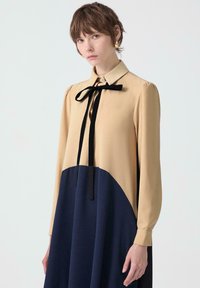 Femeie cu păr scurt brunet, purtând o rochie bej și bleumarin, cu un papion negru și cercei aurii, stând pe un fundal alb.