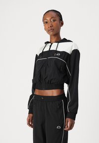 Cazadora negra con detalles blancos, diseño corto, capucha ajustable y puños elásticos. Presenta bolsillos laterales y logotipo.