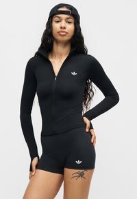 Femme portant un haut noir à manches longues zippé et un short assorti avec des logos Adidas, casquette noire portée à l’envers, et tatouage d’araignée sur la cuisse gauche.