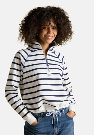 Frau mit lockigem Haar lächelt, trägt einen weißen Pullover mit marineblauen horizontalen Streifen und halbem Reißverschluss, kombiniert mit blauen Jeans mit Kordelzugbund.