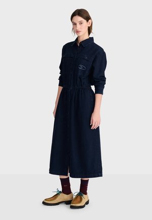 Jeune femme debout portant une longue robe bleu marine boutonnée à manches longues, des chaussettes bordeaux et des chaussures beiges à lacets.