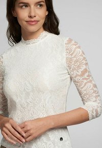 Weißes Spitze-Top mit hohem Kragen, 3/4-Ärmeln, floralen Mustern und einem dekorativen Knopf auf Taillenhöhe. Texturiertes Design überall.