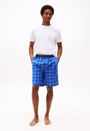 Pantaloni corti a quadretti blu con una cintura elastica, caratterizzati da un logo nero "TOMMY HILFIGER", abbinati a una t-shirt bianca a maniche corte.