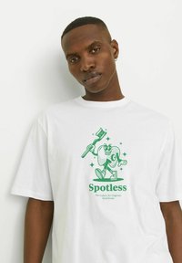 Tricou alb din bumbac cu un grafic verde ce prezintă un personaj zâmbitor cu o periuță de dinți, textul "Fără pete" se află dedesubtul designului.