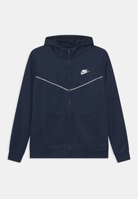 Marinblå zip hoodie tillverkad av slät tyg. Har en huva, två framfickor, vit Nike-logotyp och en reflekterande rand över bröstet.