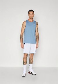 Muž s tetovaniami, ktorý má na sebe svetlomodré bezrukávové tričko, biele športové šortky, biele ponožky a biele tenisky, stojí pred jednoduchým pozadím.