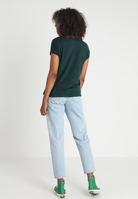 Mörkgrön kortärmad t-shirt med avslappnad passform, kombinerad med ljusblå raka jeans och gröna högskaftade sneakers på en enfärgad bakgrund.