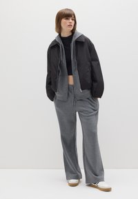 Schwarze Bomberjacke über einem grauen Hoodie, kombiniert mit grauen Jogginghosen. Das Outfit hat eine lässige Passform und elegante weiße Sneaker mit braunen Sohlen.