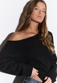 Črn kratek pulover z odkritimi rameni in rebrastimi rokavi ter robom, izdelan iz mehke pletene tkanine, kombiniran z teksturiranimi jeansi iz jeansa.