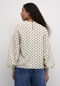 Crème blouse met polka dots in zwarte stippen, voorzien van lange pofmouwen, een keyhole achterkant en elastische manchetten. Gemaakt in combinatie met blauwe denim.