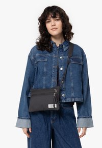 Černá crossbody taška s texturovaným povrchem, vybavená zipovým zavíráním a nastavitelným popruhem. Model nosí modrou džínsovou bundu a kalhoty.