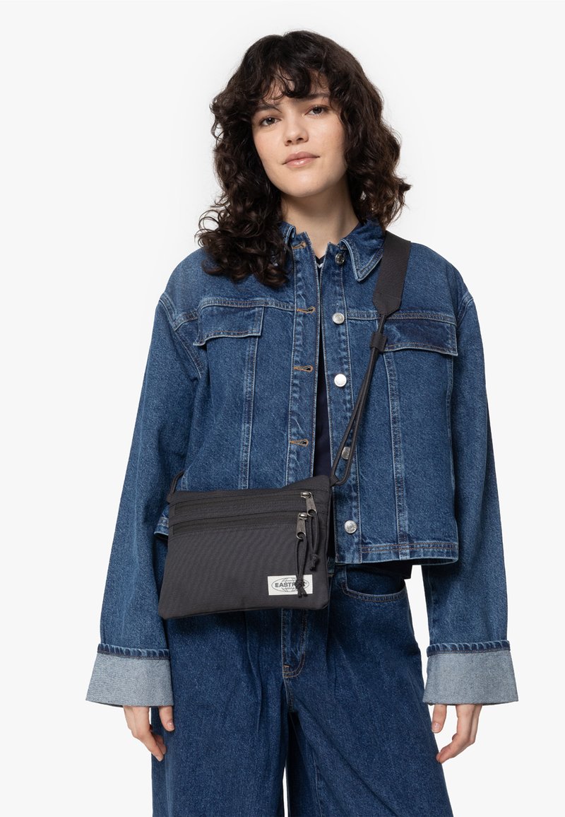 Černá crossbody taška s texturovaným povrchem, vybavená zipovým zavíráním a nastavitelným popruhem. Model nosí modrou džínsovou bundu a kalhoty.