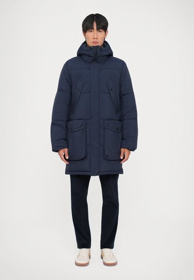 Chaqueta de plumas azul marino con capucha, de corte oversized, con dos grandes bolsillos frontales y cierre de cremallera. Textura suave, diseño acolchado.